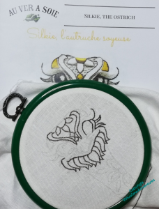 Cover of the instructions for silkie the ostrich showing behind a green flexihoop with part of the design outlines stitched