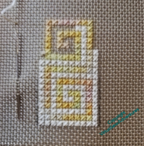 Close up of a piece of canvaswork in cream and yellow. There is a needle in the canvas beside the worked piece, which is about as high as the needle.