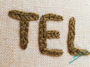 Close up of three letters, "T", "E", "L" worked in a mystery stitch, in an olive-brown colour on sandy fabric.