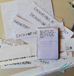 A desk, covered in slips of paper mentioning "Excavations" and "Akhetaten", topped by a notebook with a quotation from the book "Nefertiti Lived Here" scrawled in it.