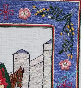 Close up of the top right corner of the border. The background is trellis couching in several shades of blue, the edges are yellow, red and green, and the corner motif shows sprigs of broom intertwined with dog roses. The rose stem used a fawn thread.