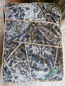 Top-down view of a footstool. The cover is made of many pieces of black and gold figured upholstery velvet, with gold piping.