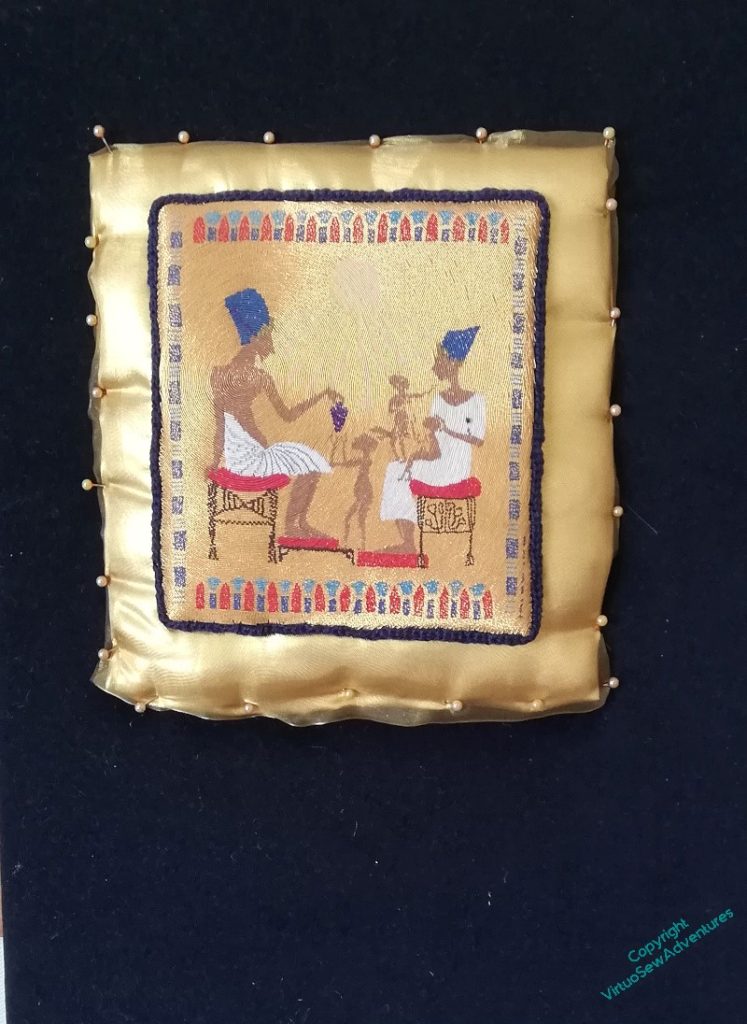 Photograph of the or nué of the Amarna Family Group presented over a gold cushion against the navy stela