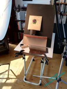 An easel of sorts is set up at the focus of photographic equipment. The Violets in Stumpwork are on a small pad on that easel.