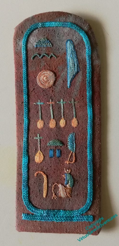 An embroidered version of the Cartouche of Nefertiti, in turquoise and yellow on a brown marbled fabric.