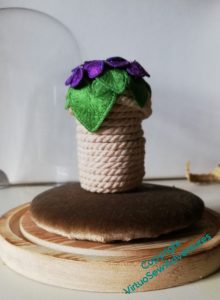 Coiled pot with violets attached, set upon the cushion, with the glass dome in the background.