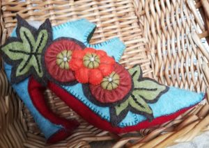 Side view of a high-heeled shoe made of felt and stuffed, then decorated with felt flowers. The shoe has a red sole, like the famous Louboutins.