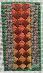 A panel of needlepoint, mosaic stitch in green around the edge, three columns of grey on either side, and then a panel down the middle in brown and flame colours, with green bosses in the gaps.